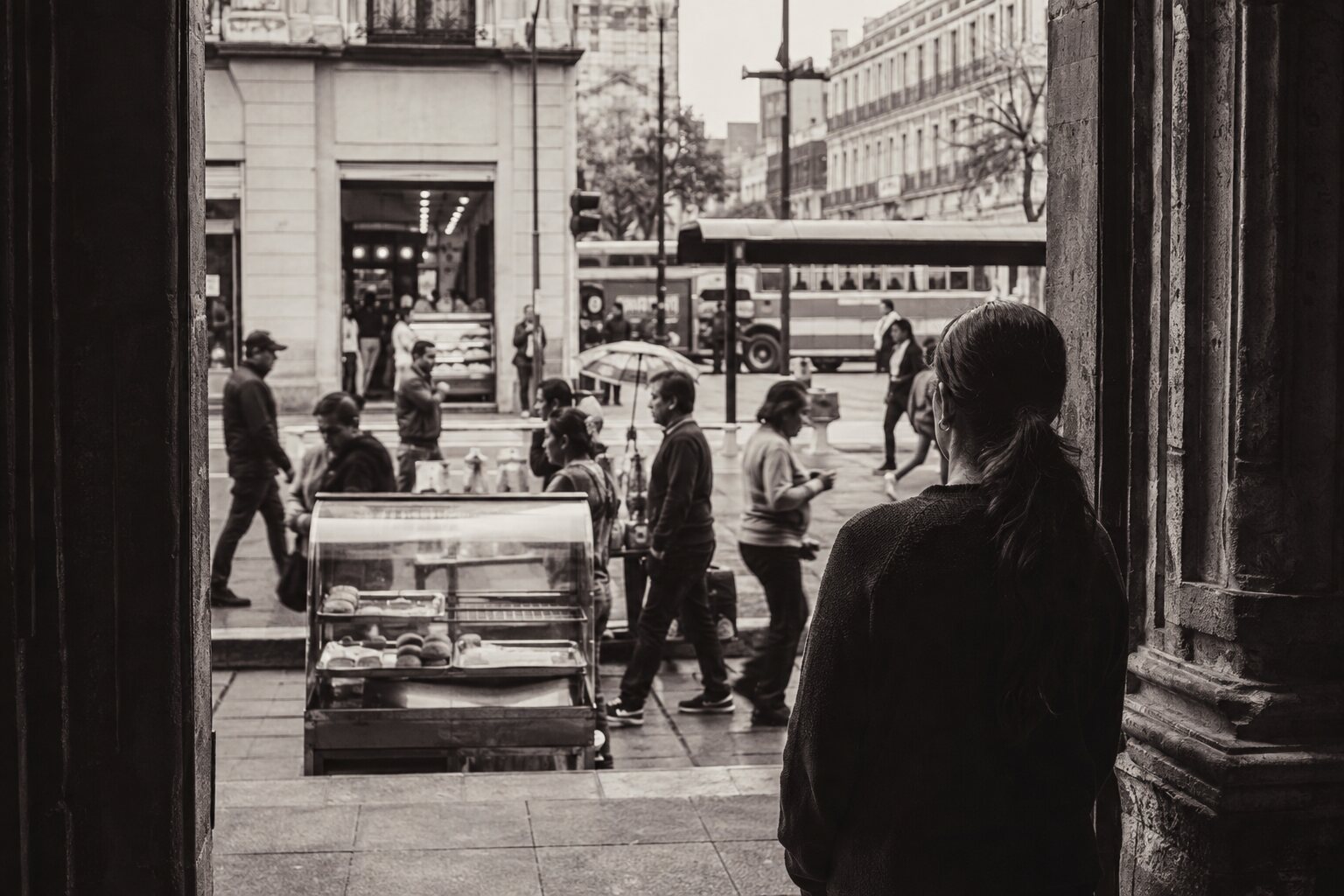 Mujer observando la actividad cotidiana de una ciudad latinoamericana desde un umbral.